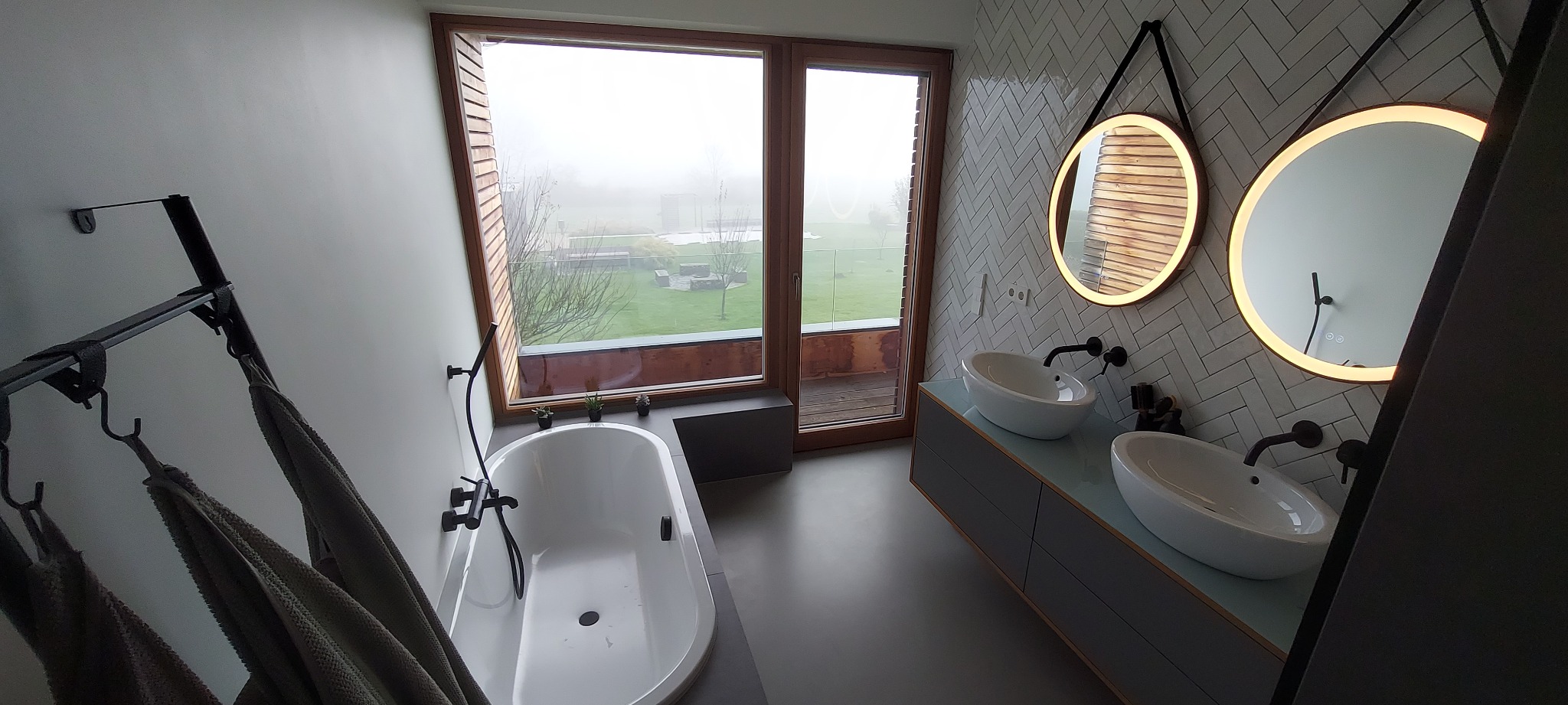 Nowoczesna łazienka z wanną przy oknie z widokiem na zewnątrz, dwie okrągłe umywalki z podświetlanymi lustrami i białe płytki ułożone w jodełkę.