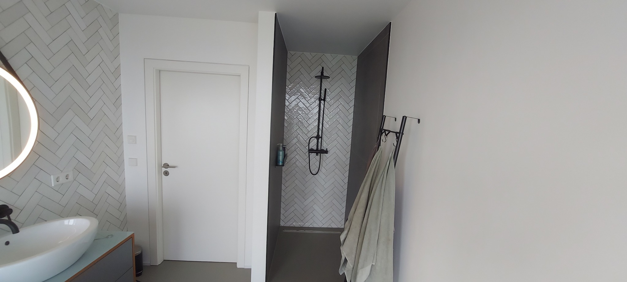 Nowoczesna łazienka z kabiną prysznicową typu walk-in, wykończona białą, ułożoną w jodełkę glazurą. Widoczna umywalka z okrągłym lustrem z podświetleniem LED.