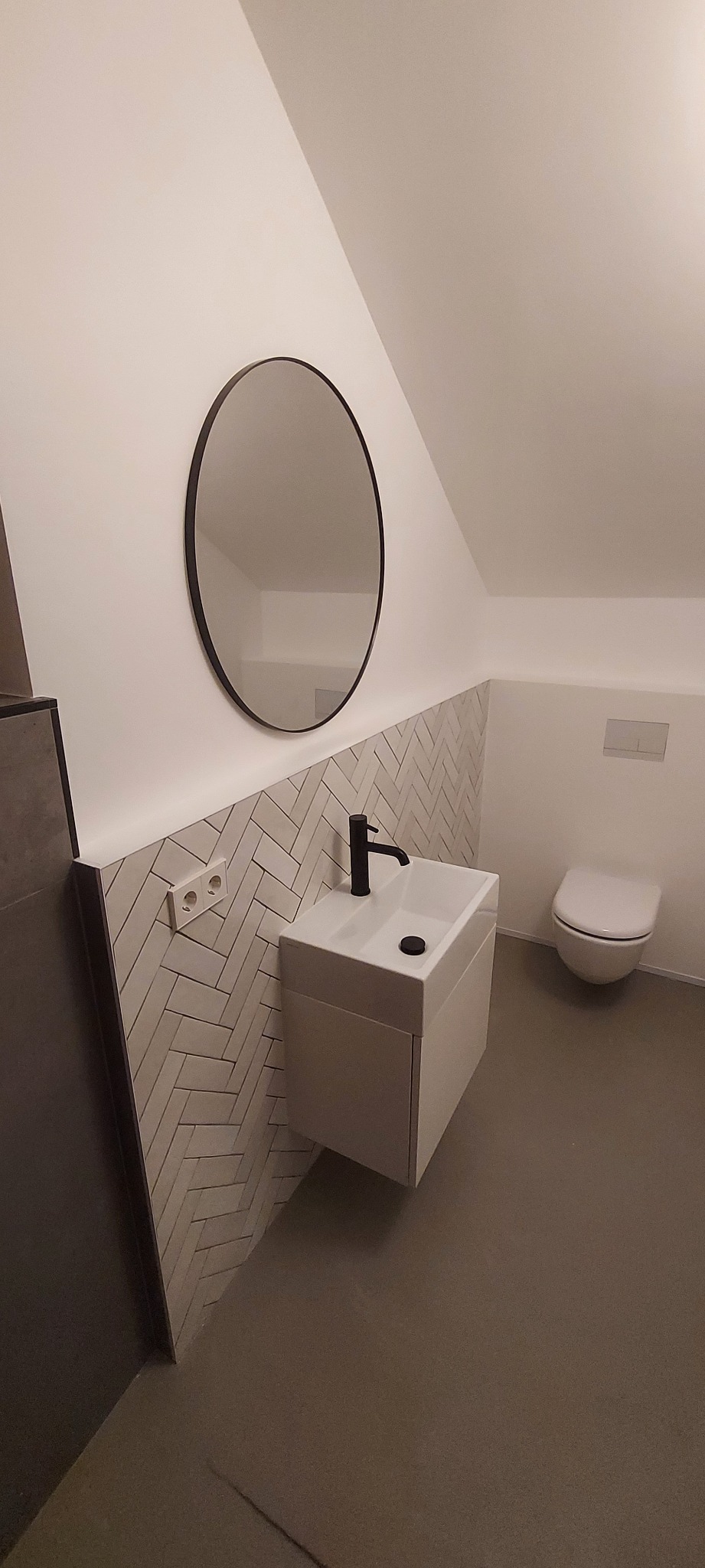 Nowoczesna łazienka na poddaszu z umywalką, lustrem w czarnej ramie i płytkami ułożonymi w jodełkę. Minimalistyczny design, stonowana kolorystyka, widok na toaletę.