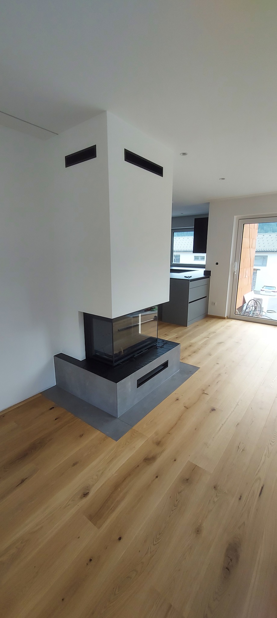 Salon z drewnianą podłogą i nowoczesnym kominkiem z przeszkloną obudową. W tle widoczna kuchnia i okno z widokiem na zewnątrz.