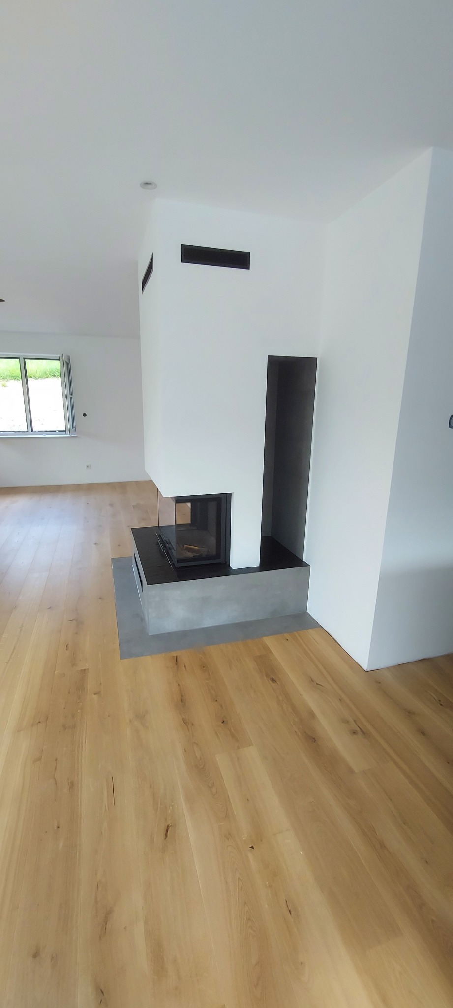 Nowoczesny kominek z szarą obudową w jasnym wnętrzu. Parkiet z naturalnego drewna, białe ściany i okno w tle. Minimalistyczny design.