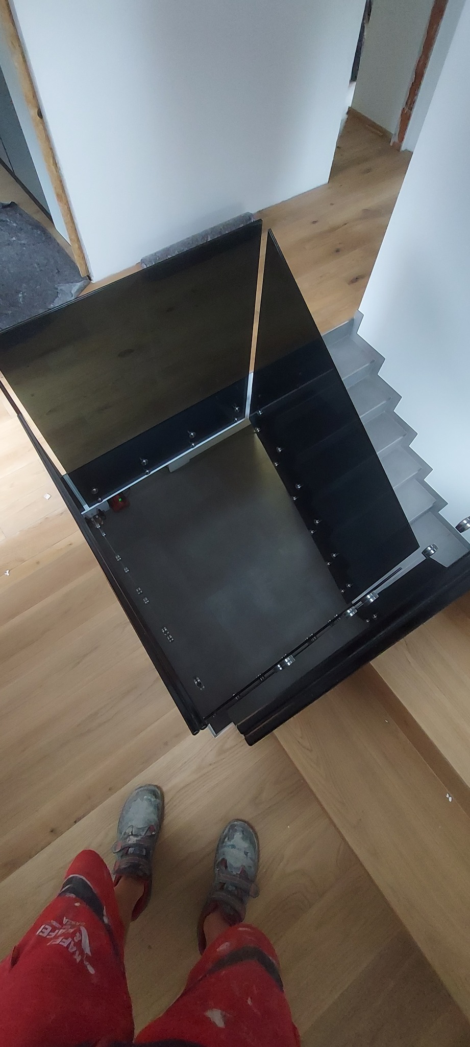 Nowoczesna balustrada schodowa z czarnego szkła, zamontowana na drewnianych schodach. Widoczne detale mocowań i odbicia światła. Ujęcie z góry, z widocznymi butami montera.