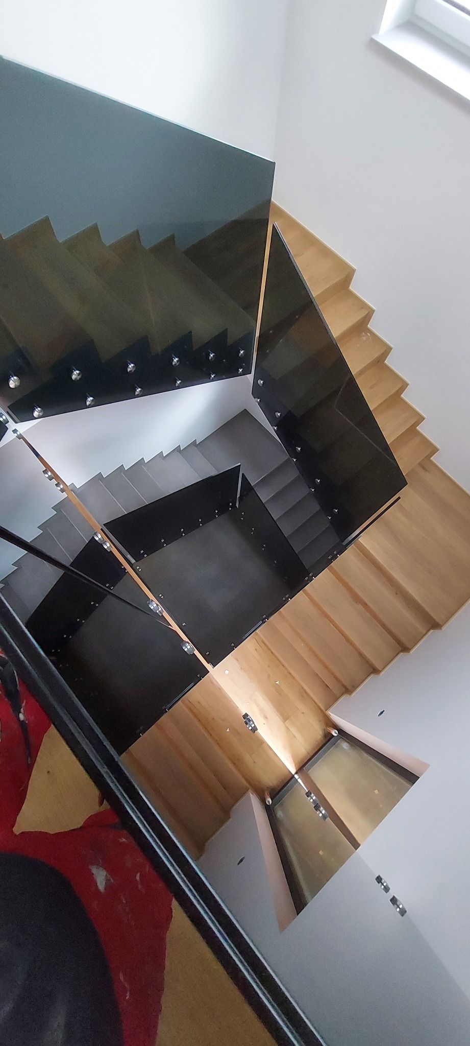 Nowoczesne schody z drewnianymi stopniami i szklaną balustradą, widok z góry. Nietypowy układ schodów z elementami czerni i szarości. W tle fragment obrazu.