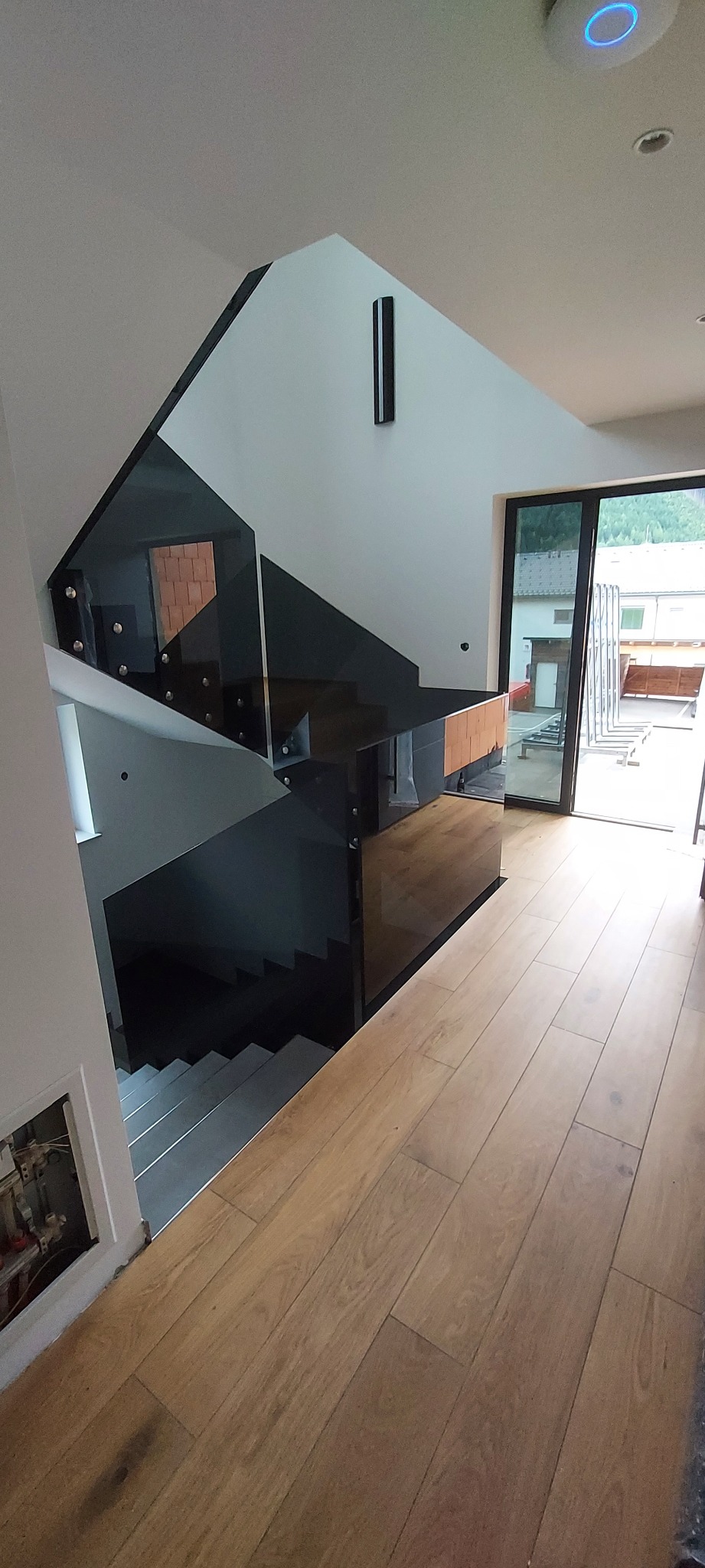 Nowoczesne schody z czarnymi szklanymi balustradami i drewnianymi stopniami, widok z góry na jasną podłogę i przeszklone drzwi balkonowe.