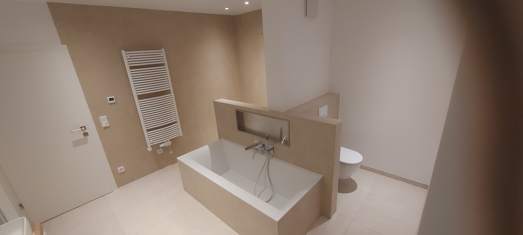 Nowoczesna łazienka w beżach: wanna z zabudową, grzejnik drabinkowy, toaleta. Minimalistyczny design, jasne płytki, punktowe oświetlenie. Wykończenie wnętrz.