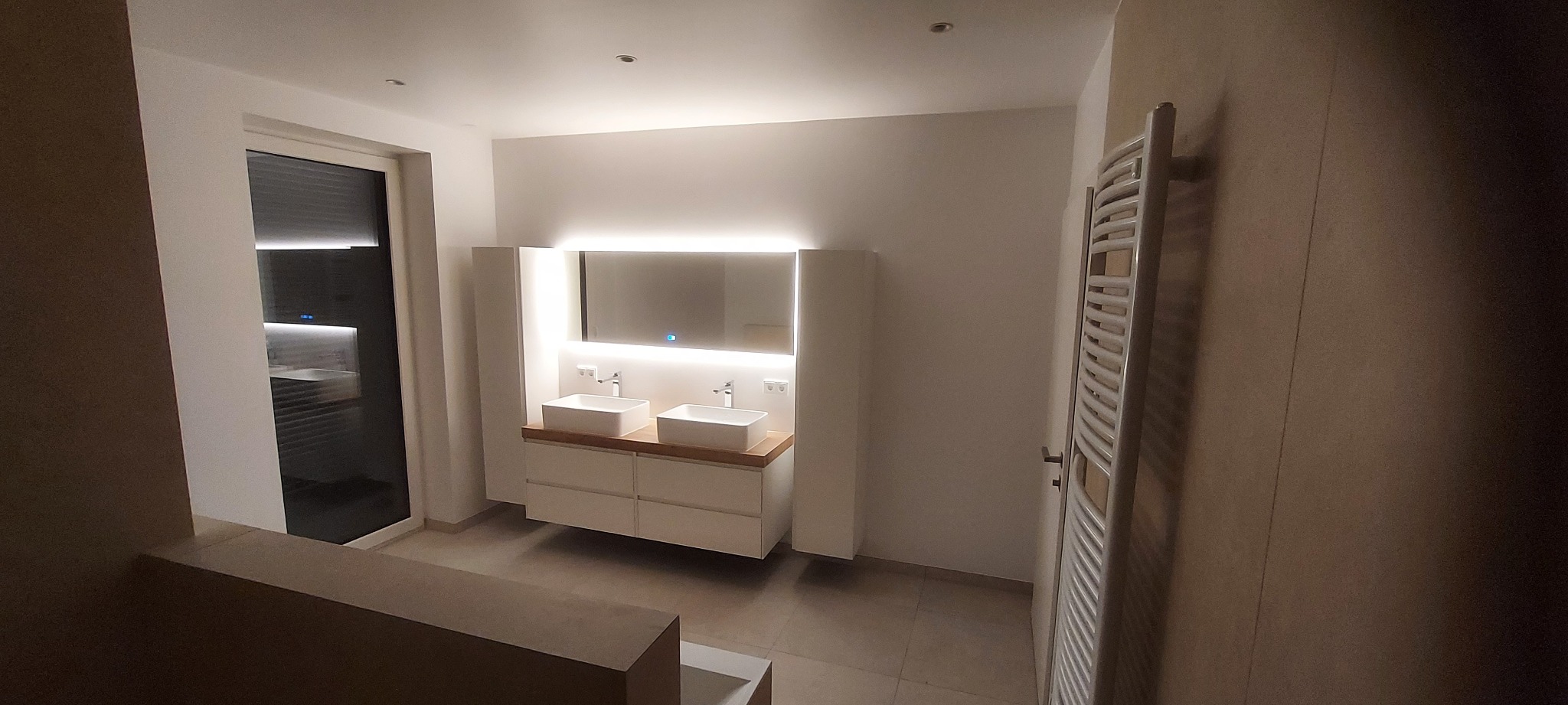 Nowoczesna łazienka z podświetlanym lustrem, dwiema umywalkami nablatowymi i grzejnikiem drabinkowym. Minimalistyczny design, jasne kolory i subtelne oświetlenie.