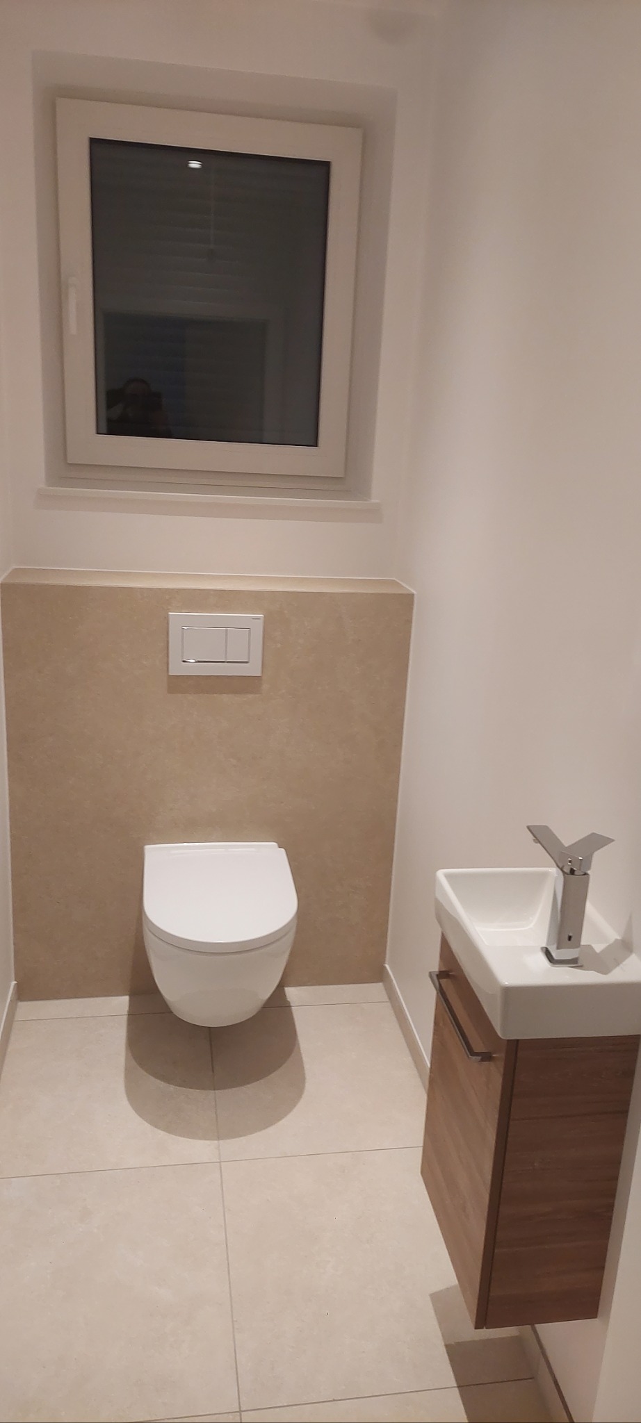 Nowoczesna, minimalistyczna toaleta z podwieszaną miską WC, umywalką z szafką i beżowymi płytkami podłogowymi. Widoczne okno z białymi ramami.