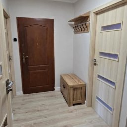 MD-BUD - Jasne panele podłogowe w przedpokoju z drewnianą skrzynią, wieszakiem i dwojgiem drzwi w odcieniach brązu. Okrągła lampa sufitowa.