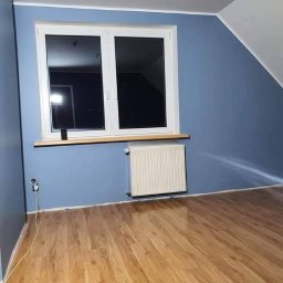 MD-BUD - Pokój na poddaszu po malowaniu ścian na niebiesko. Widoczne okno, grzejnik i drewniana podłoga. Ściany pomalowane na jednolity kolor, bez smug.