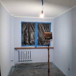 MD-BUD - Pokój po malowaniu na jasny odcień niebieskiego, zabezpieczone okno folią, grzejnik pomalowany na biało, podłoga zabezpieczona folią, lampa na statywie.