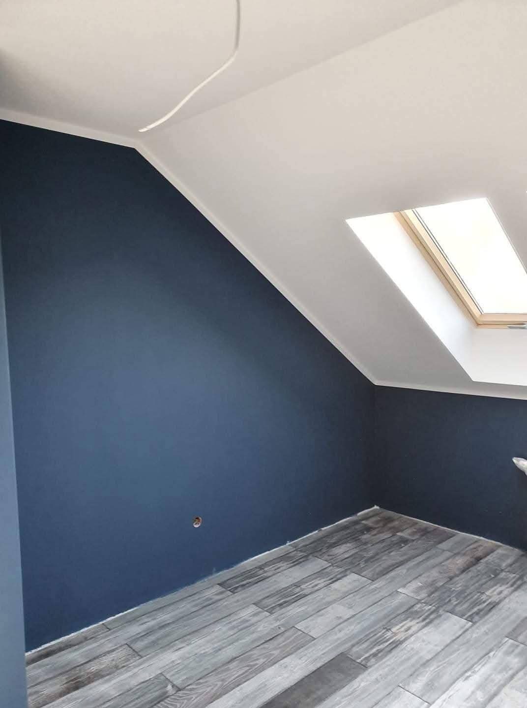 Pomieszczenie na poddaszu po malowaniu ścian na granatowo i wykończeniu podłogi panelami imitującymi drewno. Widoczne okno dachowe i biały sufit.