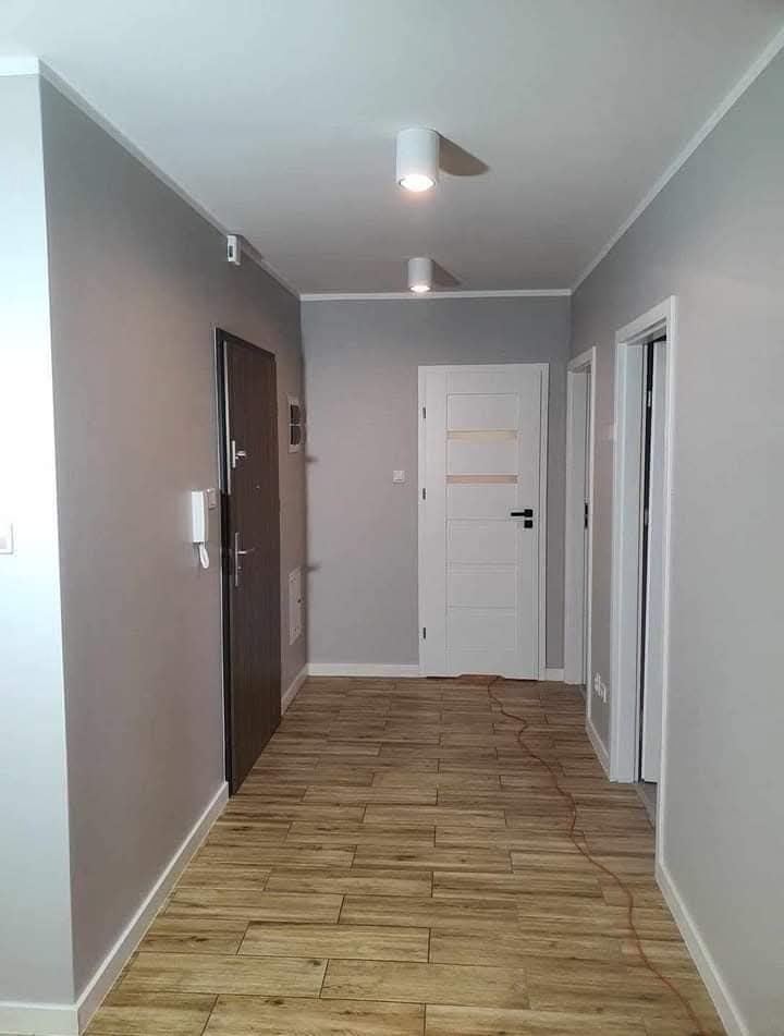 Jasne wnętrze korytarza po remoncie: szare ściany, drewnopodobna podłoga, białe drzwi i listwy przypodłogowe. Dwa nowoczesne, okrągłe plafony sufitowe.