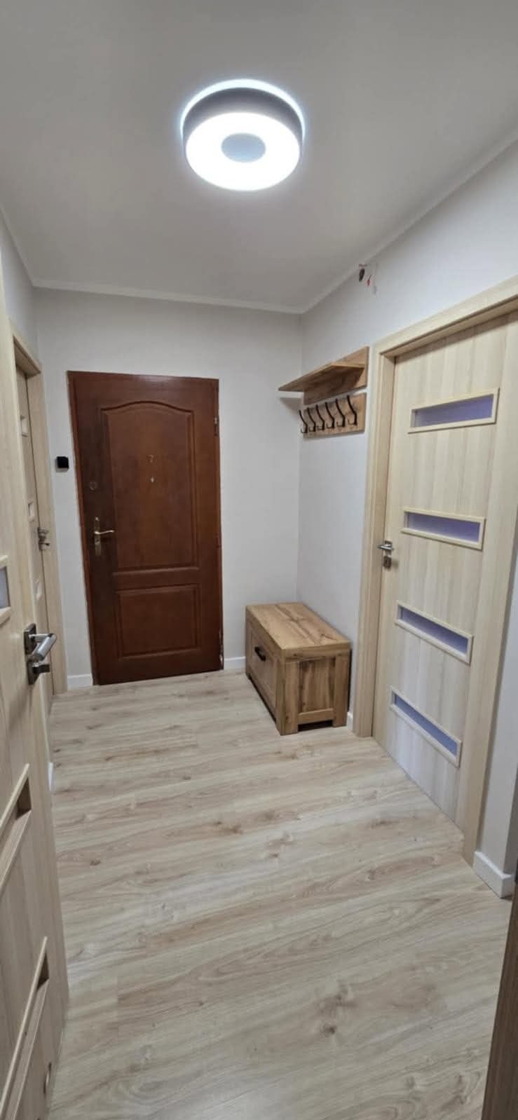 Jasne panele podłogowe w przedpokoju z drewnianą skrzynią, wieszakiem i dwojgiem drzwi w odcieniach brązu. Okrągła lampa sufitowa.