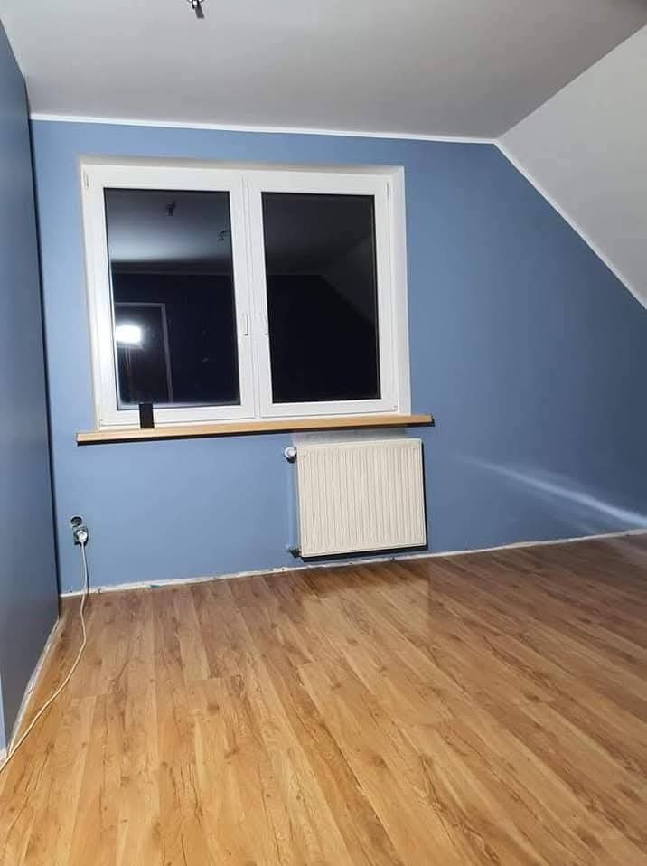 Pokój na poddaszu po malowaniu ścian na niebiesko. Widoczne okno, grzejnik i drewniana podłoga. Ściany pomalowane na jednolity kolor, bez smug.