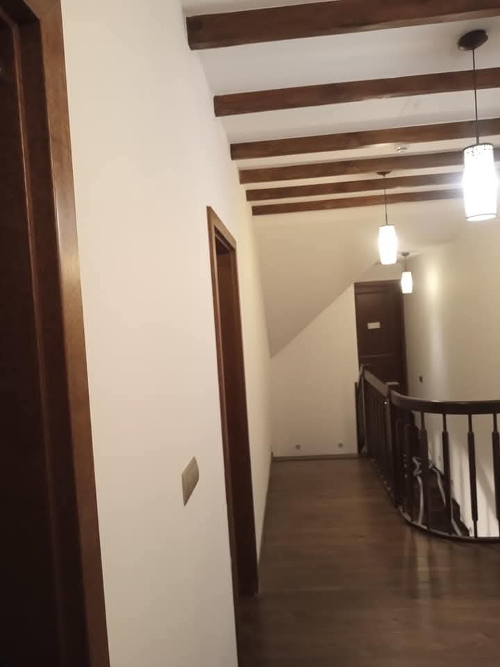 Korytarz z drewnianą podłogą i belkami stropowymi. Ściany w jasnym odcieniu, oświetlenie punktowe. Widoczne drzwi z drewnianymi futrynami i balustrada.