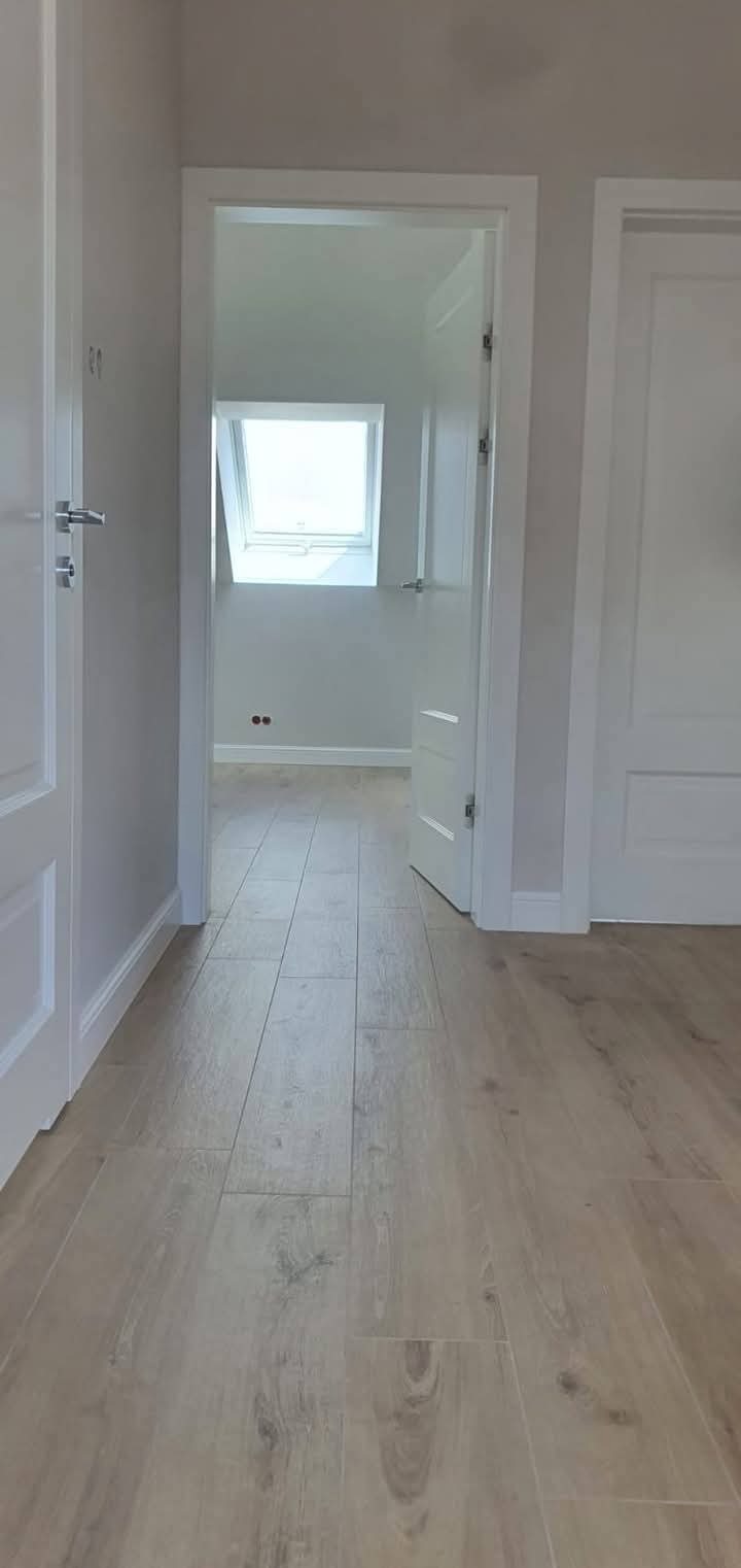 Jasne panele podłogowe w nowym pokoju z widokiem na okno dachowe przez otwarte drzwi. Świeżo pomalowane ściany w odcieniach szarości i białe listwy przypodłogowe.