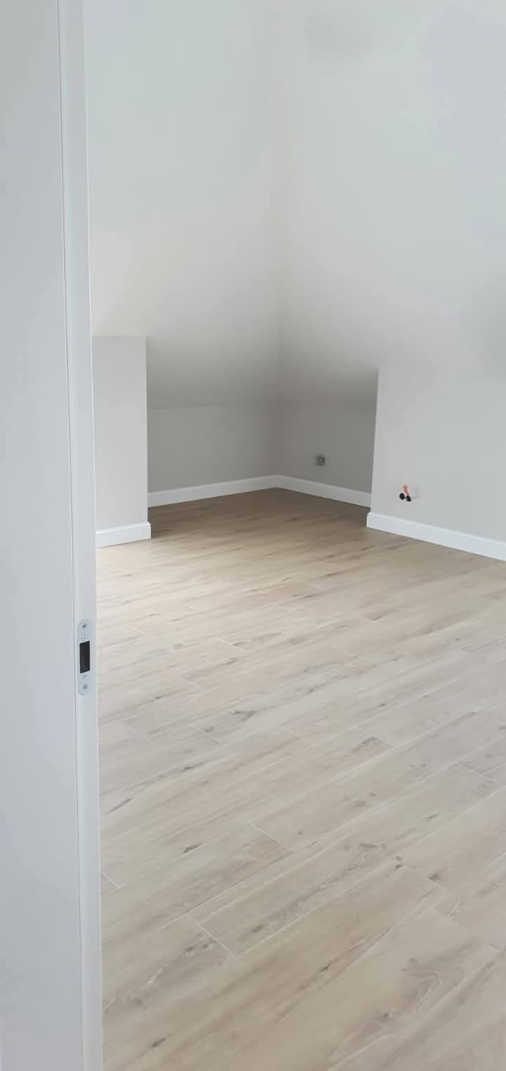 Jasne panele podłogowe imitujące drewno w nowo wykończonym pokoju na poddaszu. Ściany pomalowane na szaro, białe listwy przypodłogowe i ościeżnica drzwi.