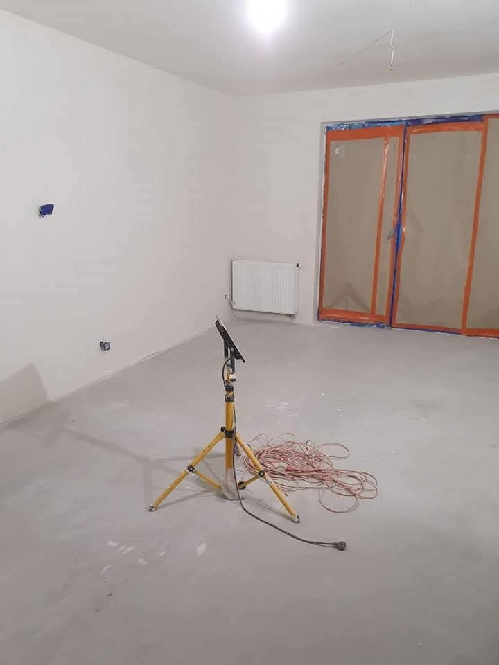 Wnętrze po szpachlowaniu w Jerzmanowej: gładkie, białe ściany, zabezpieczone okna folią, lampa budowlana na trójnogu i zwinięty kabel na podłodze.