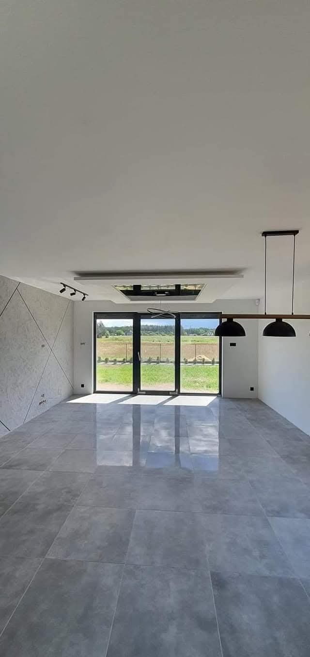 Wykończone wnętrze przestronnego salonu z szarą, błyszczącą podłogą z płytek, dekoracyjną ścianą z geometrycznym wzorem i dużym oknem tarasowym z widokiem na zieleń.