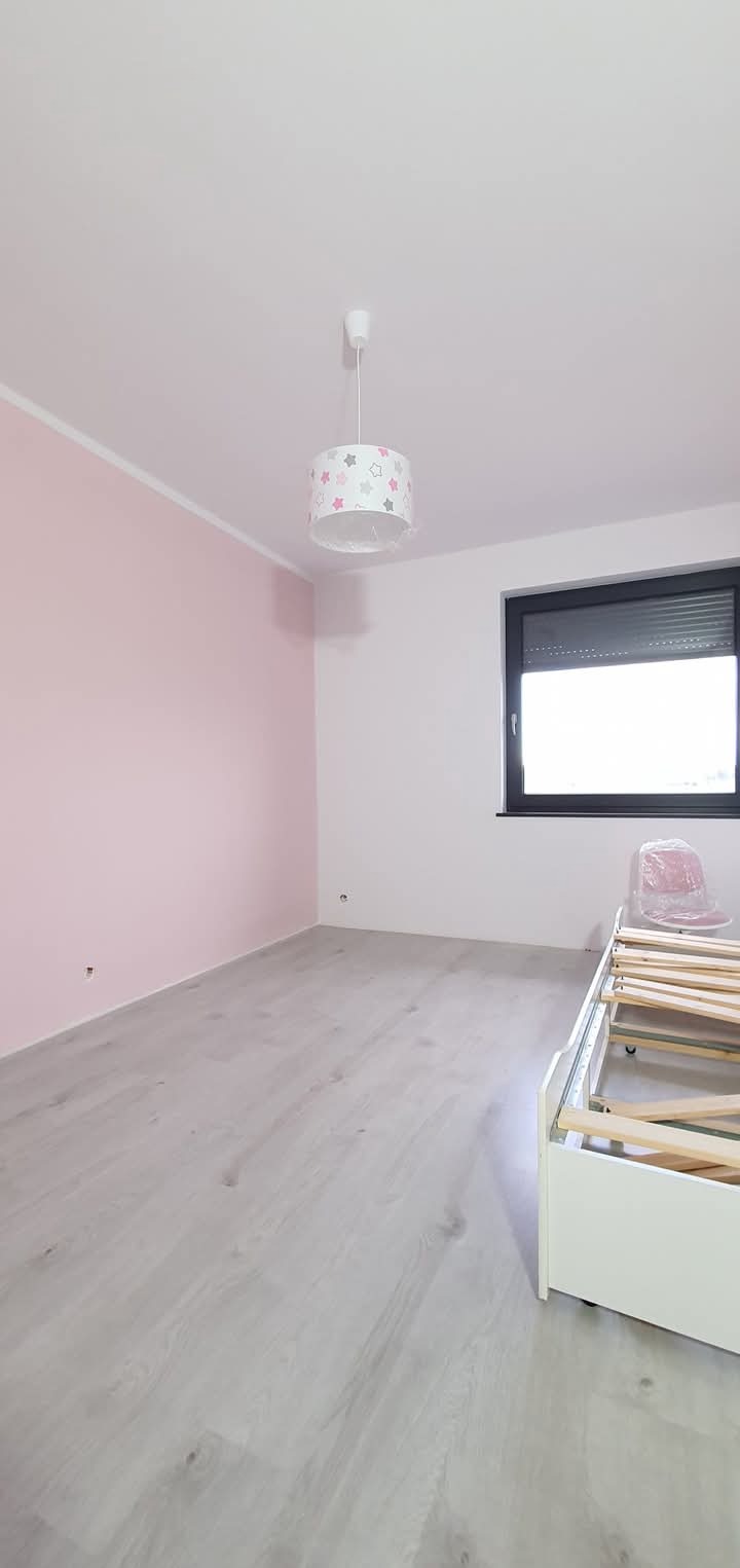 Pokój po malowaniu: różowa ściana, biały sufit, szara podłoga, okno z roletą i lampa ze wzorem gwiazd. Częściowo widoczny biały stelaż łóżka.