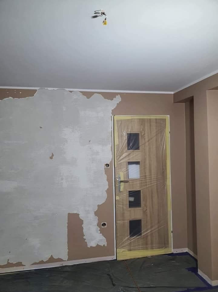 Pokój w trakcie szpachlowania: ściana z nałożoną gładzią, zabezpieczone drzwi folią malarską, widoczne puszki elektryczne. Przygotowanie do malowania.