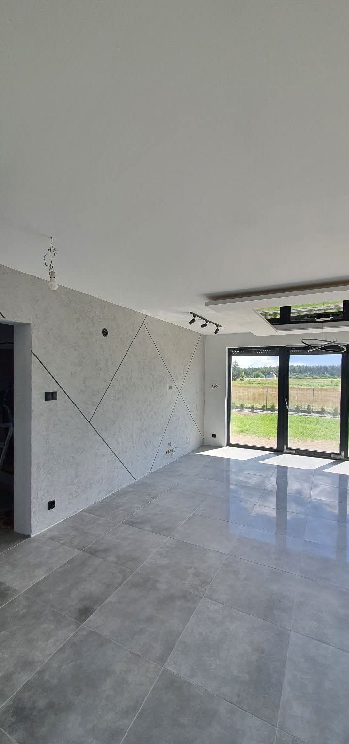 Nowoczesne wnętrze z szarą, geometryczną ścianą i błyszczącą podłogą z płytek. Widoczne okno z widokiem na zewnątrz i minimalistyczne oświetlenie sufitowe.