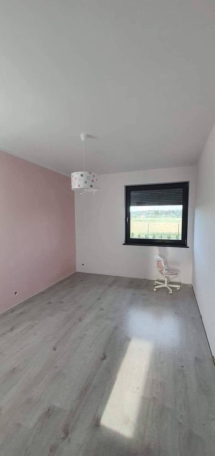 Jasne panele podłogowe i ściany po malowaniu w pokoju z oknem i krzesłem. Jedna ściana pomalowana na różowo, sufit biały, lampa z motywem gwiazd.