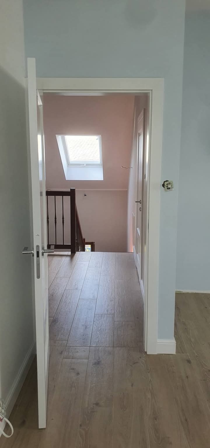 Jasne wnętrze po remoncie: drewnopodobna podłoga, białe drzwi i ościeżnice, błękitne ściany. Widok na schody i okno dachowe. Minimalistyczny design.