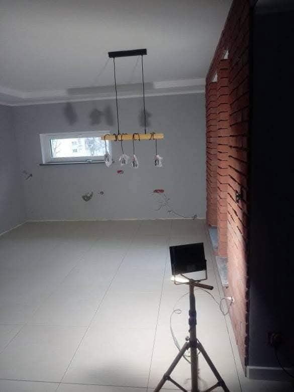 Wnętrze w trakcie wykańczania: szara ściana, okno, lampa wisząca, fragment ściany z cegły, biała podłoga z płytek i lampa budowlana na statywie.