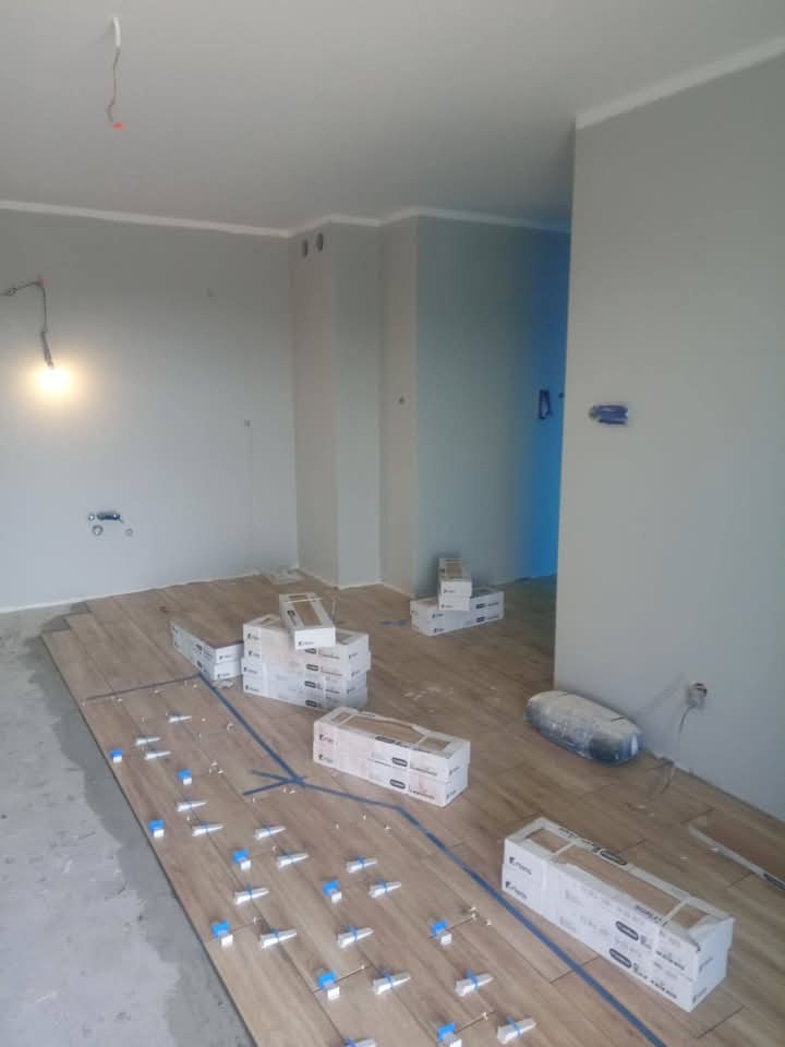 Układanie drewnopodobnych płytek podłogowych z użyciem systemu poziomowania. Częściowo wykończone, szare ściany i białe listwy przypodłogowe. Pudełka z płytkami w tle.