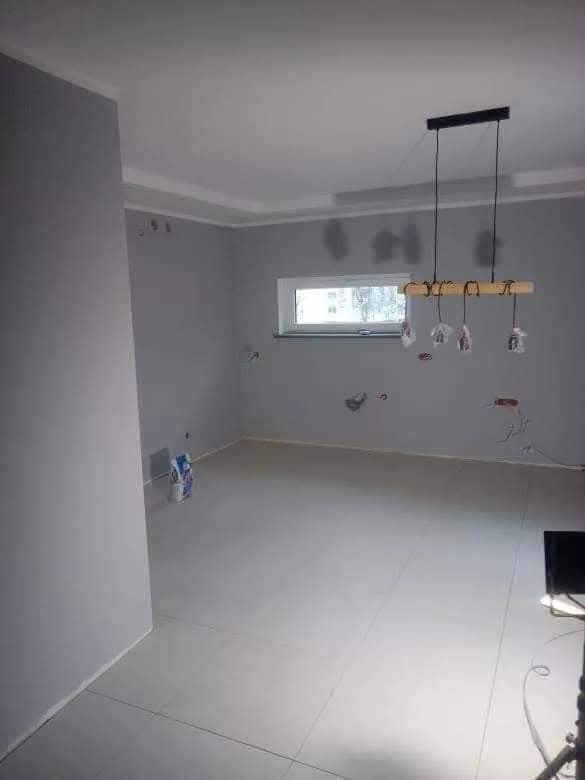 Szare wnętrze po remoncie: świeżo pomalowane ściany, ułożone płytki podłogowe, nowoczesna lampa wisząca. Widoczne ślady po instalacji elektrycznej.