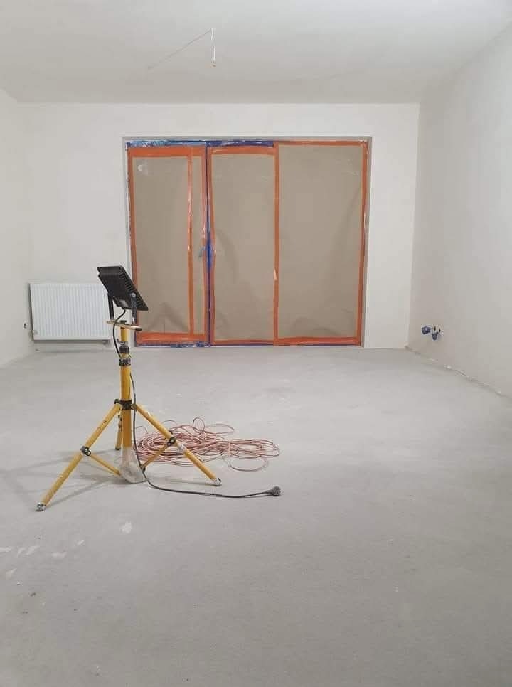 Przygotowane do malowania wnętrze z zabezpieczonymi drzwiami folią i lampą na statywie. Surowy beton na podłodze, ściany po szpachlowaniu, gotowe do nałożenia farby.