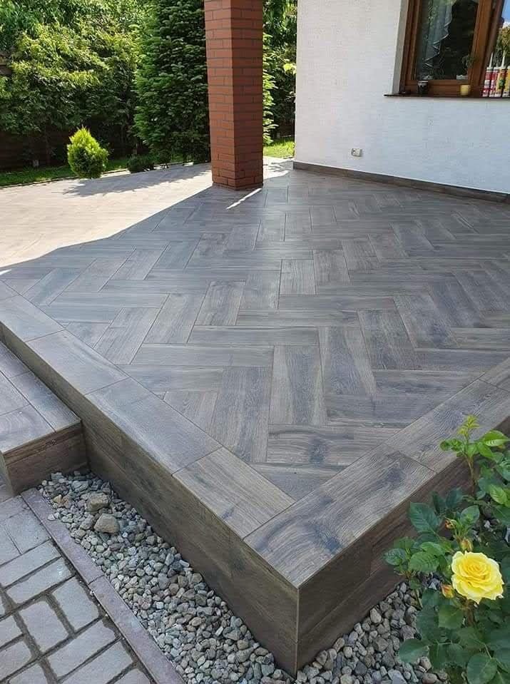 Wykończony taras z szarych płytek ułożonych w jodełkę, z obrzeżem i schodami. Widoczne kamienie dekoracyjne i fragment roślinności. Estetyczne wykończenie zewnętrzne.