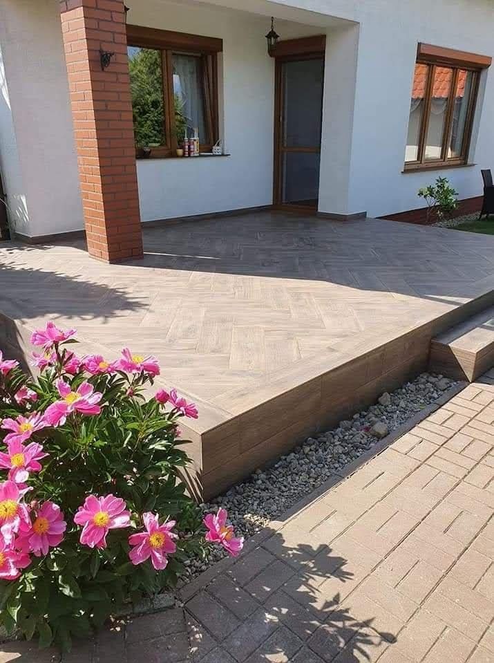 Wykończony taras z jasnobrązowych płytek ułożonych w jodełkę, z niskim murkiem i kamieniami ozdobnymi, na tle białego domu z oknami w brązowych ramach.