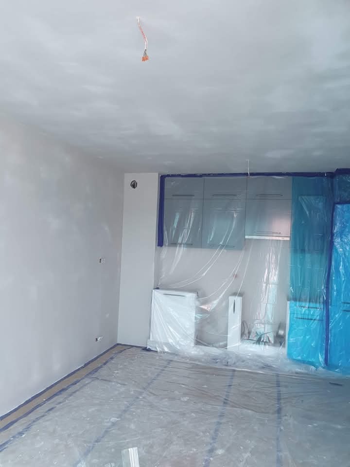 Przygotowanie do malowania: zabezpieczona folią kuchnia z szarymi szafkami, świeżo wyszpachlowane ściany, taśmy malarskie na podłodze i wystające kable z sufitu.