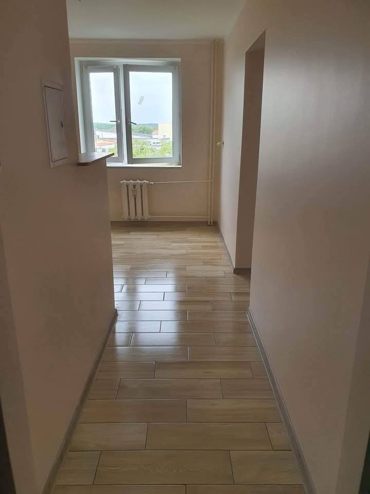 Jasne wnętrze po remoncie: świeżo pomalowane ściany w odcieniach beżu, drewnopodobna podłoga z płytek, okno z widokiem na zieleń. Minimalistyczny grzejnik.