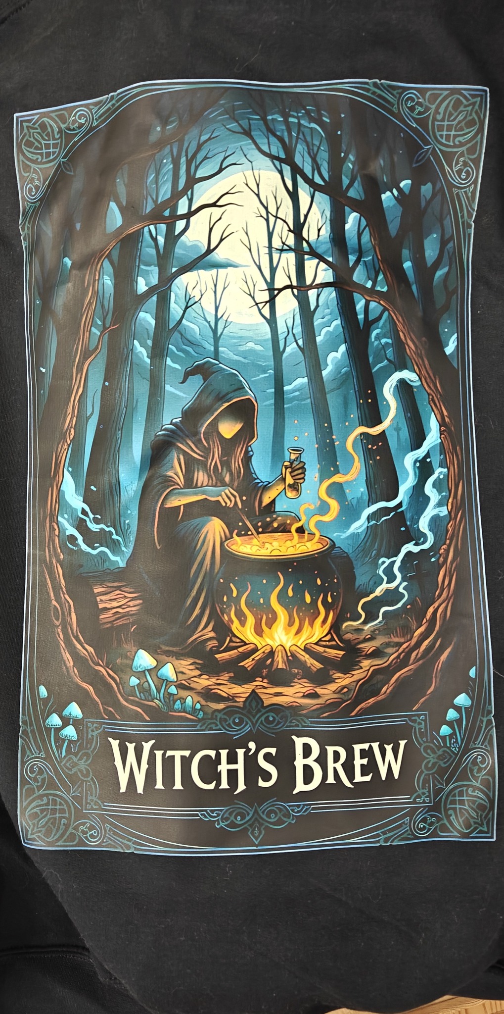 Grafika na czarnej tkaninie: czarownica warzy eliksir w lesie, kocioł z ogniem, napis 'Witch's Brew', dekoracyjne obramowanie.
