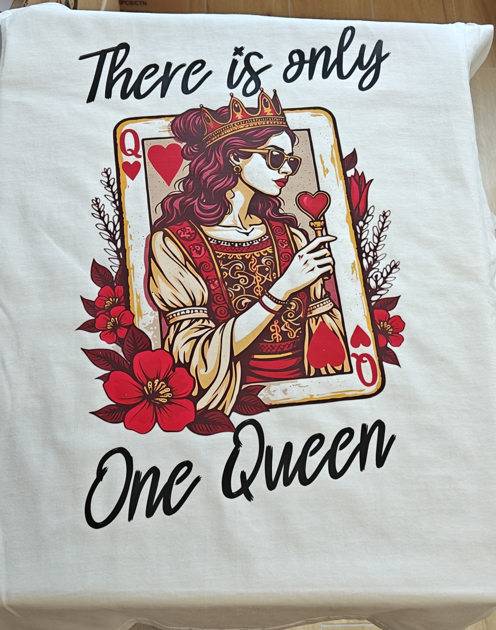 Biała koszulka z nadrukiem grafiki Królowej Kier w okularach i koronie, otoczonej kwiatami, z napisem 'There is only One Queen'.