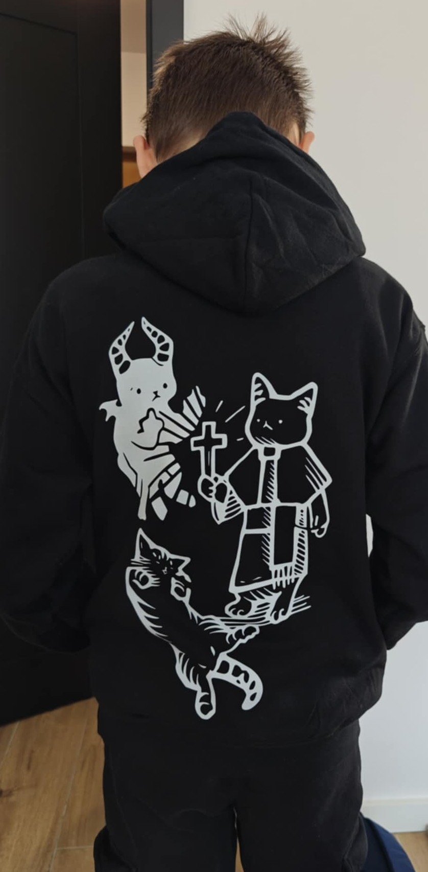 Czarna bluza z kapturem, na której widnieje biały nadruk z motywem kotów w stylu religijnym i demonicznym. Widok z tyłu na osobę noszącą bluzę.