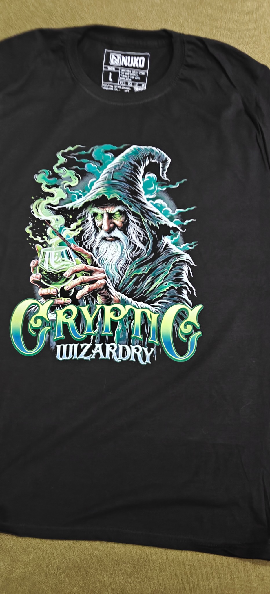 Czarna koszulka z nadrukiem czarodzieja w zieleni i błękicie, trzymającego eliksir. Napis 'Cryptic Wizardry'. Widoczna metka marki Nuko.