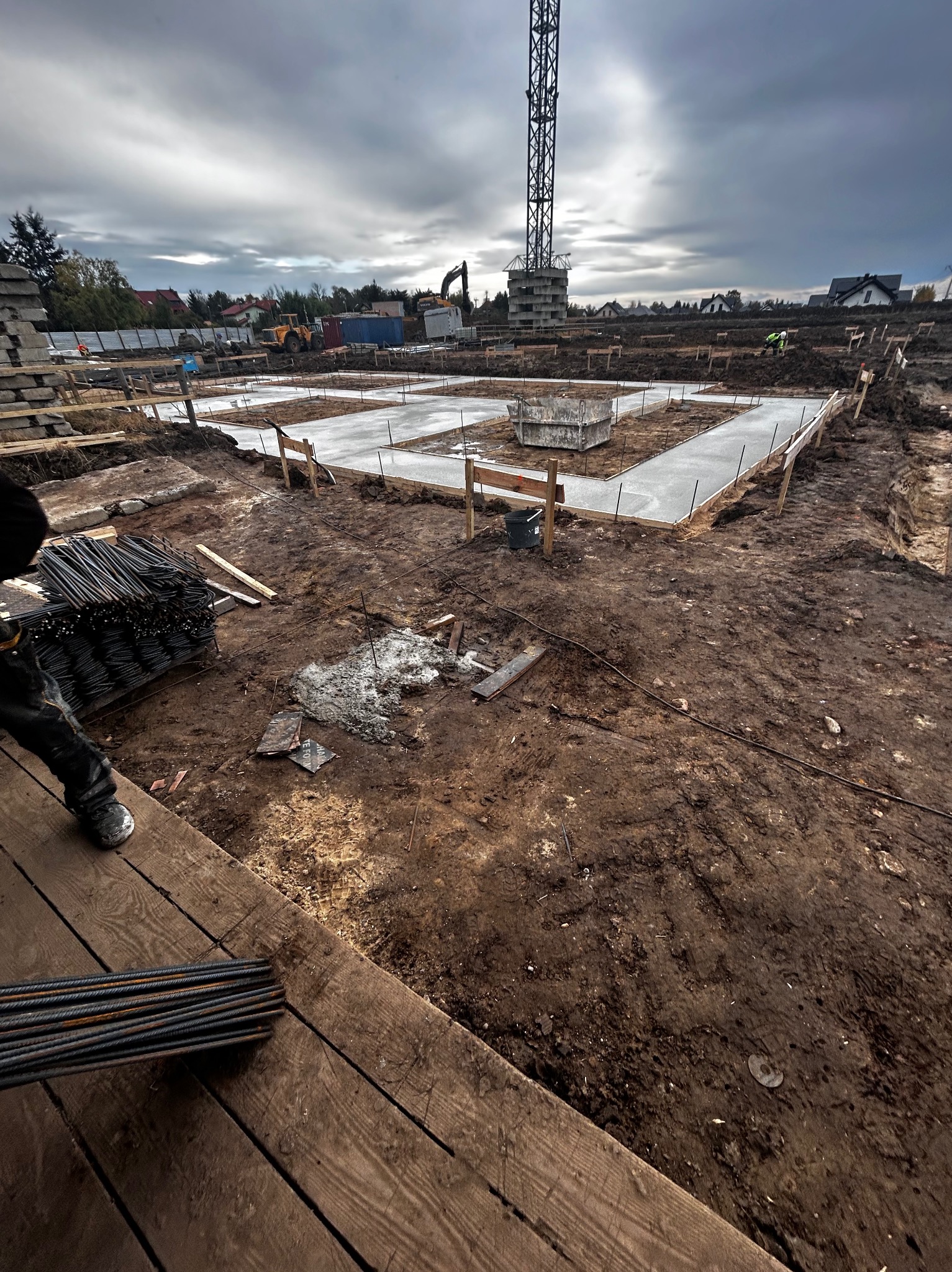 Zalane fundamenty domu jednorodzinnego, widoczne zbrojenia i szalunki. W tle dźwig budowlany i zabudowa jednorodzinna. Pochmurne niebo.