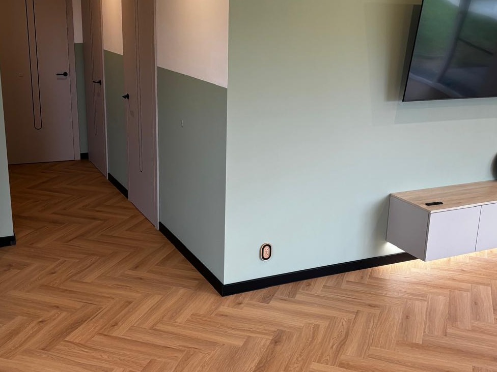 Ukończona podłoga w jodełkę z paneli, czarne listwy przypodłogowe, ściany w dwóch odcieniach zieleni, minimalistyczna szafka RTV i telewizor na ścianie.