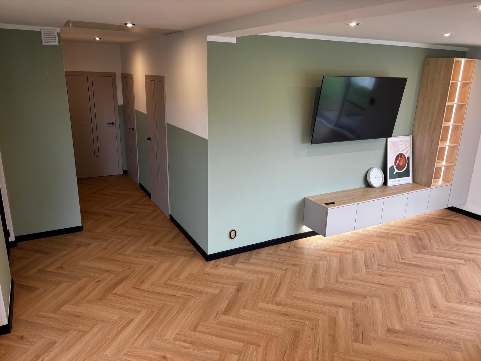 Salon z parkietem ułożonym w jodełkę francuską, ściany w odcieniach zieleni i bieli, minimalistyczne meble RTV i TV na ścianie. Nowoczesne wykończenie wnętrz.