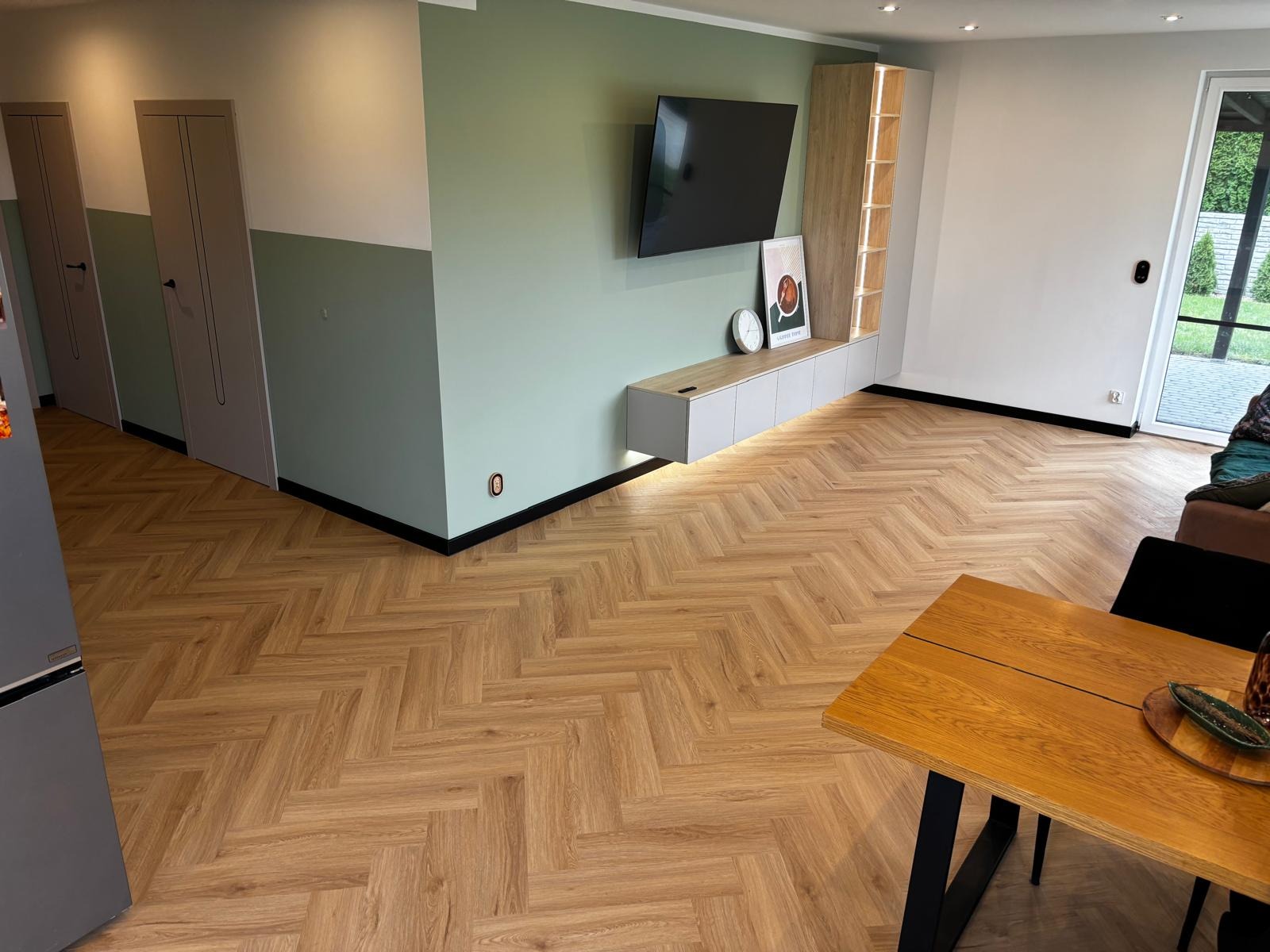 Salon z parkietem ułożonym w jodełkę francuską. Jasne panele podłogowe, nowoczesne meble, ściany w dwóch odcieniach zieleni. Widok na drzwi balkonowe i fragment stołu.