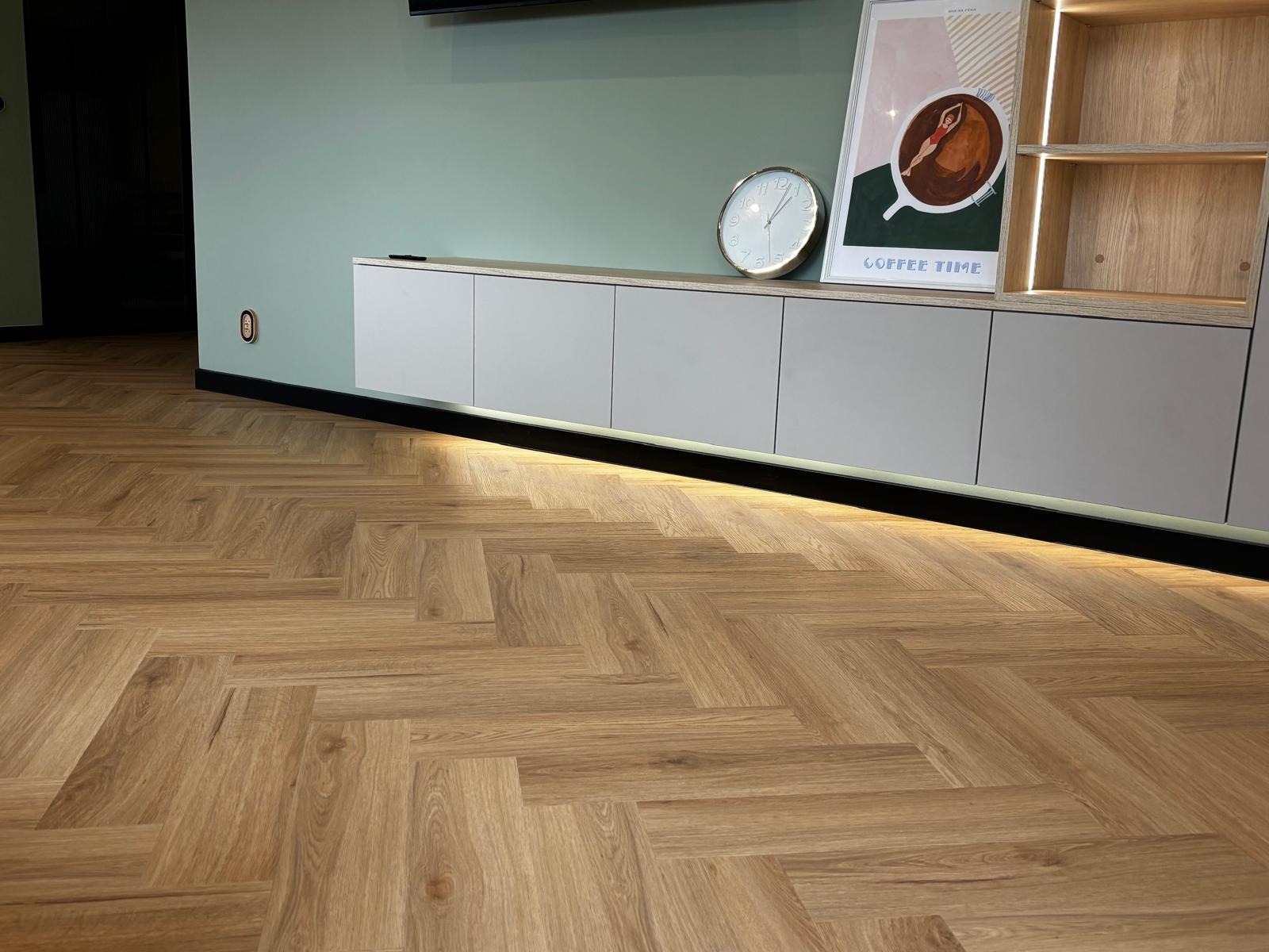 Parkiet ułożony w jodełkę francuską, oświetlenie LED pod szafką RTV, zegar i plakat z motywem kawy na tle miętowej ściany. Nowoczesne wykończenie wnętrza.