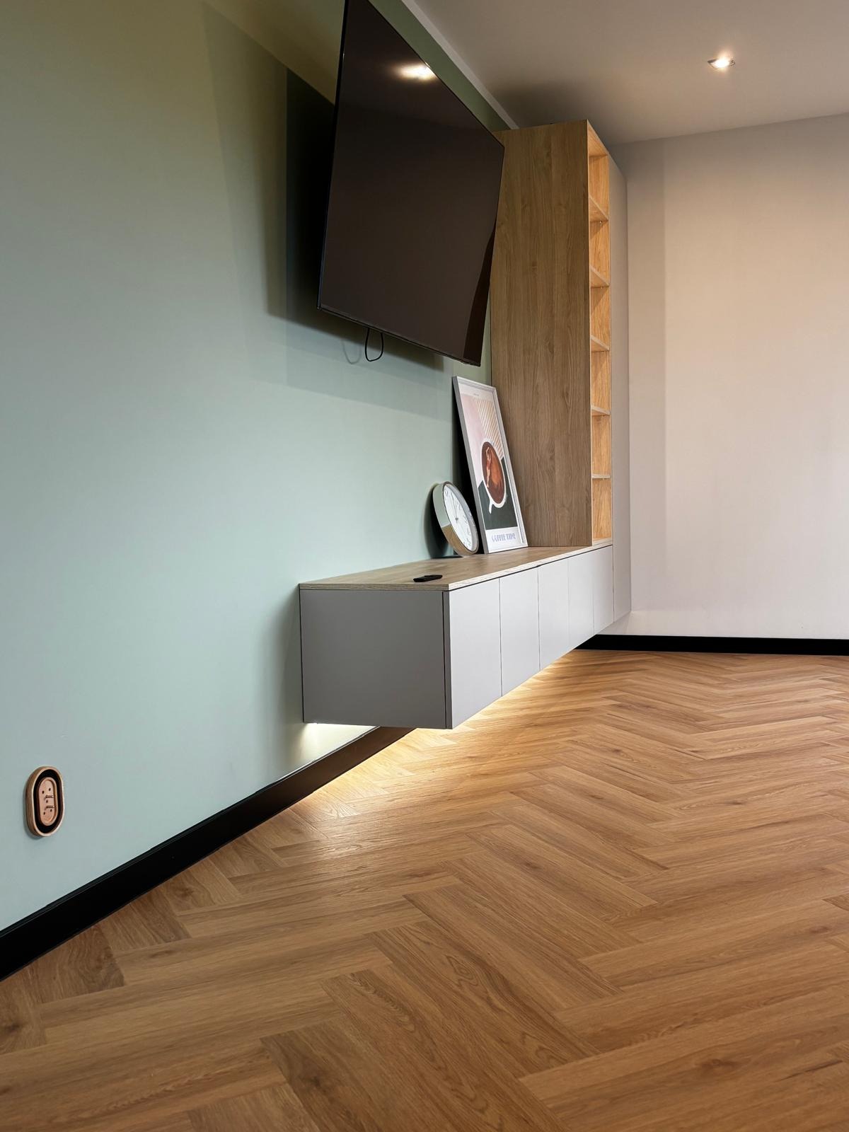 Salon z podłogą ułożoną w jodełkę, wisząca szafka RTV z podświetleniem LED, ściana w kolorze miętowym, nowoczesne gniazdko. Parkiet w ciepłym odcieniu dębu.