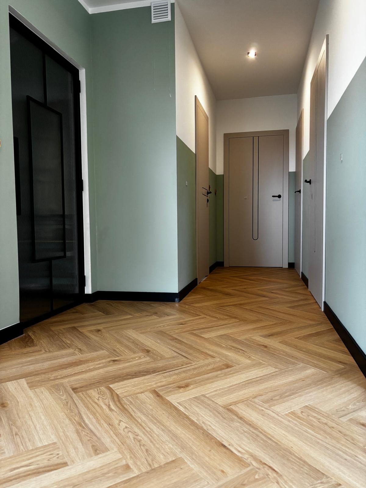Ukończony korytarz z parkietem ułożonym w jodełkę, ściany w kolorze zieleni i bieli, czarne listwy przypodłogowe i drzwi w odcieniach beżu i czerni.