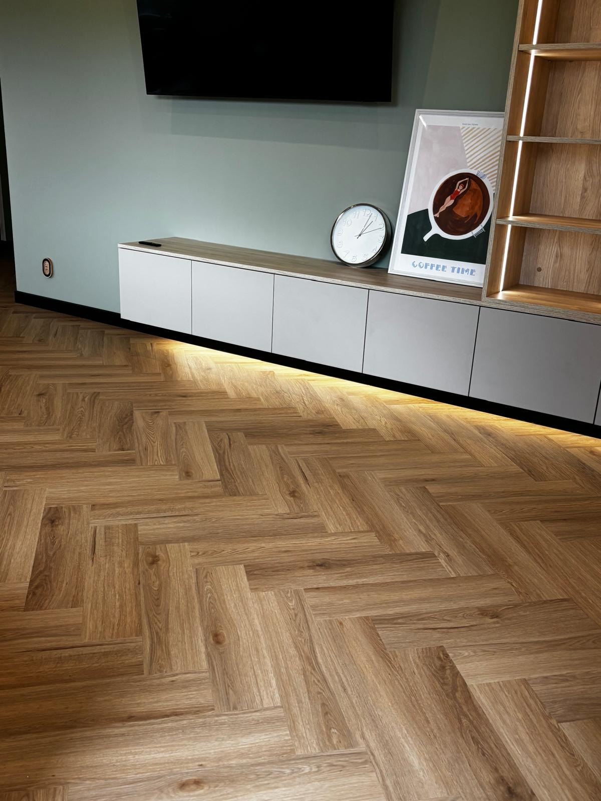 Ułożona podłoga z paneli w jodełkę, ciepłe oświetlenie LED pod szafką RTV, zegar i plakat z kawą na tle ściany w odcieniach zieleni. Nowoczesne wykończenie wnętrza.