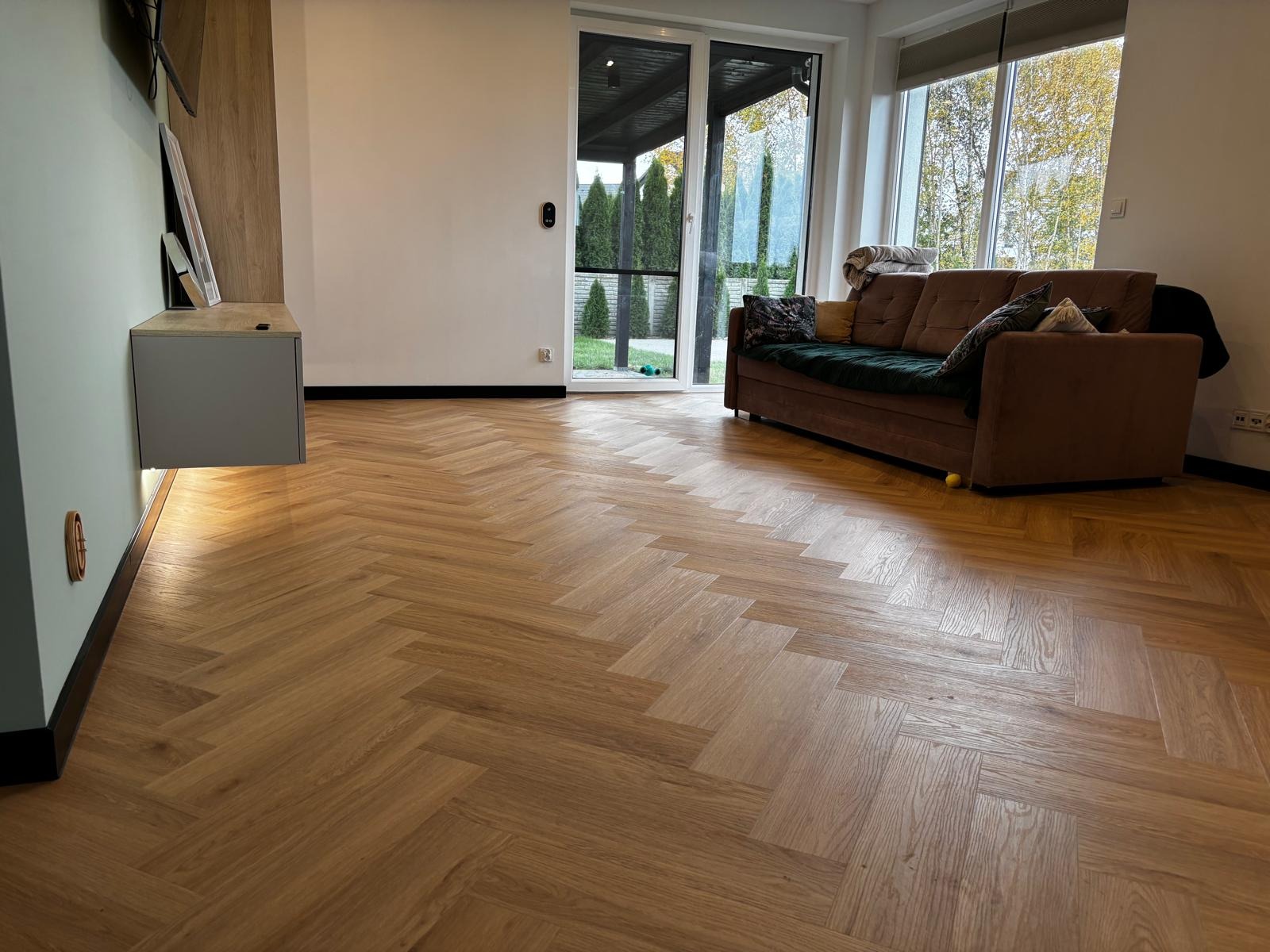 Salon z parkietem ułożonym w jodełkę francuską, widok na taras przez duże okno. Nowoczesne wnętrze z elementami drewna i zieleni. Jasne, naturalne światło.