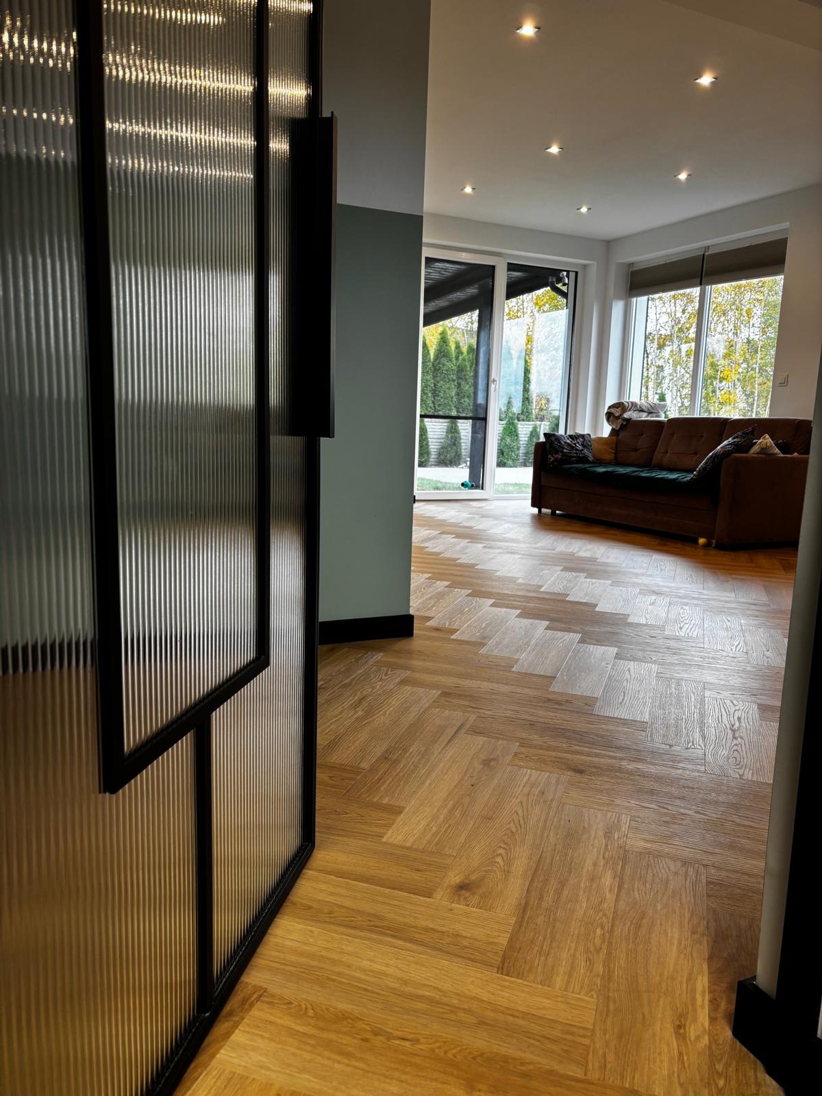 Parkiet w jodełkę francuską z jasnego drewna w nowoczesnym salonie. Widok zza szklanych drzwi z czarną ramą. W tle sofa i okno.